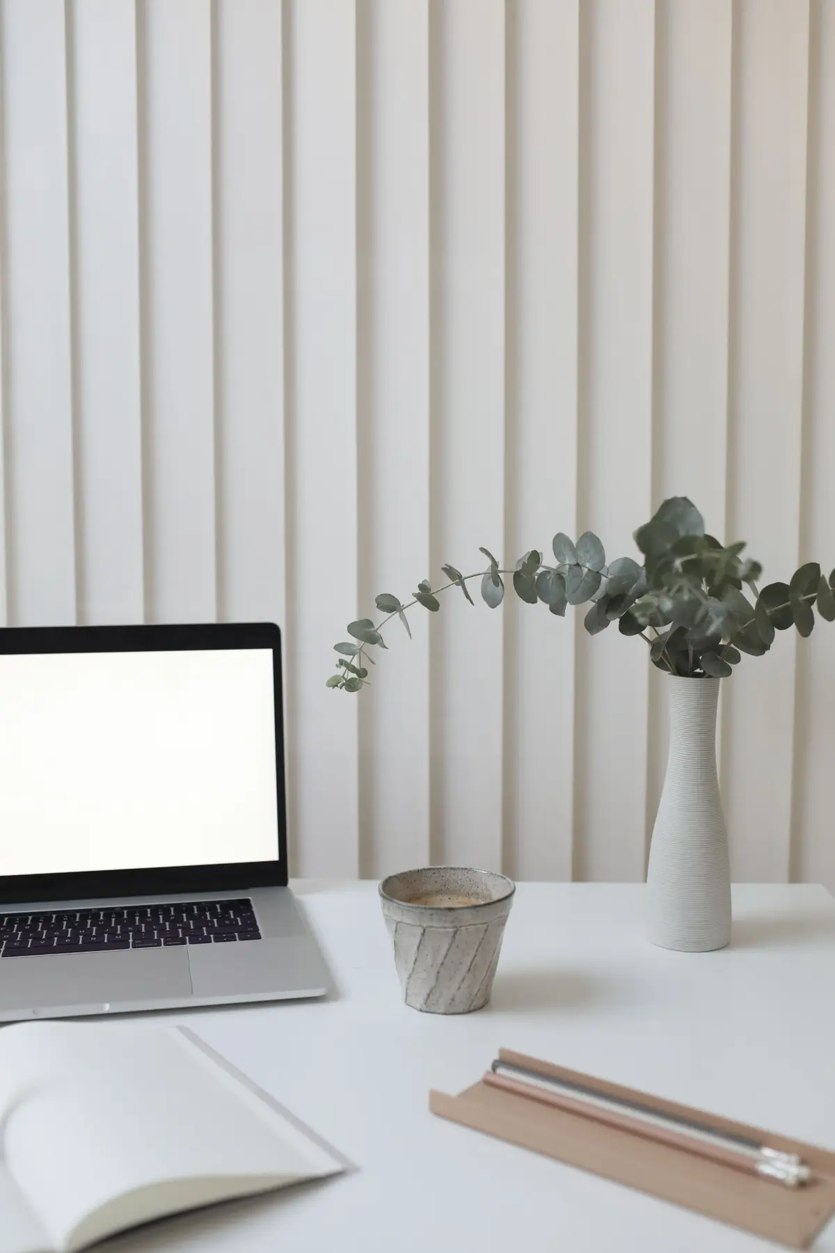 A calm desk with eucalyptus in a white vase, an open notebook and a cup of coffee